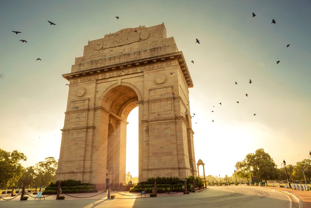 The India Gate monument in New Delhi, India, stands tall against a bright sunset sky with many birds flying.