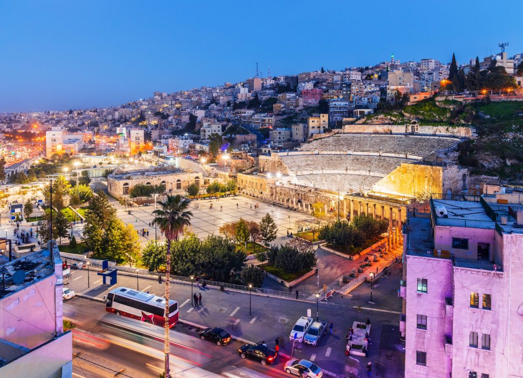 Aerial view of Amman's Roman Theatre at dusk, with city buildings and illuminated streets.