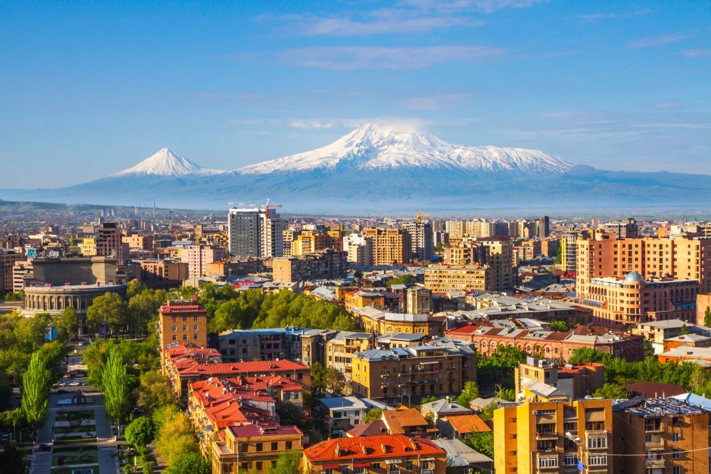 Yerevan cityscape with buildings, trees, and the snow-capped Mount Ararat in the background under a blue sky.