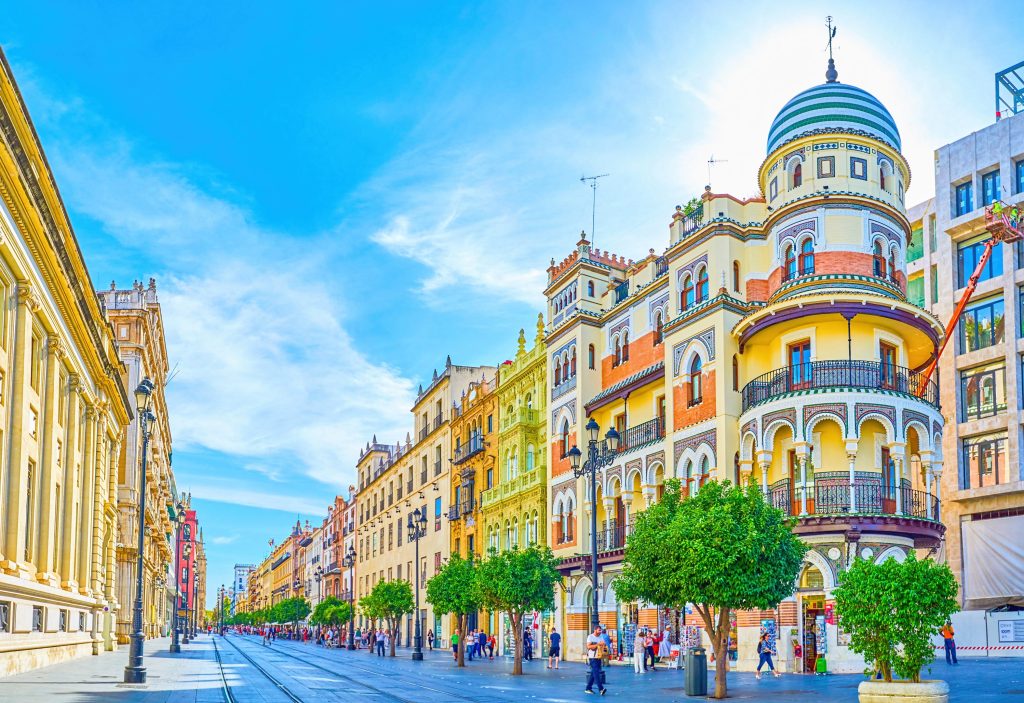 Bright Seville street view with colorful buildings, trees, and people walking under a blue sky.