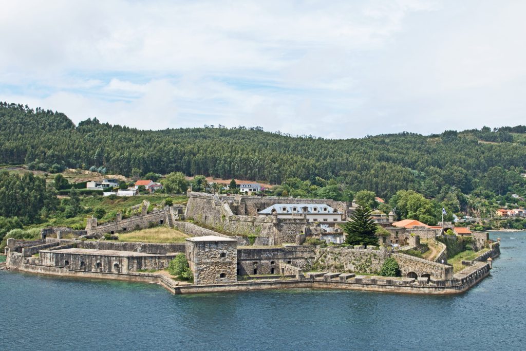 Stone fortress on a peninsula surrounded by blue water, green trees and cloudy sky.
