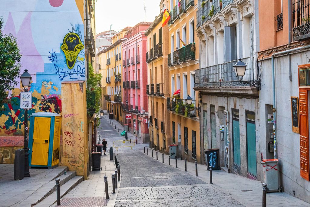 A narrow cobblestone street in Madrid, Spain, lined with colorful buildings and graffiti art.