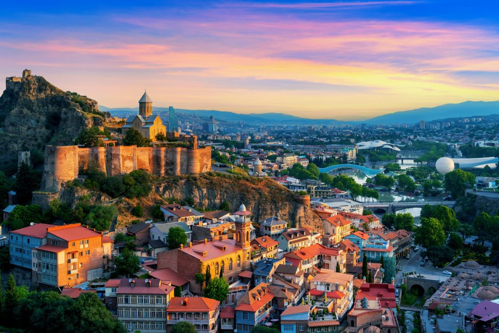 Aerial view of Tbilisi, Georgia, featuring Narikala Fortress, colorful buildings, and a vibrant sunset sky.