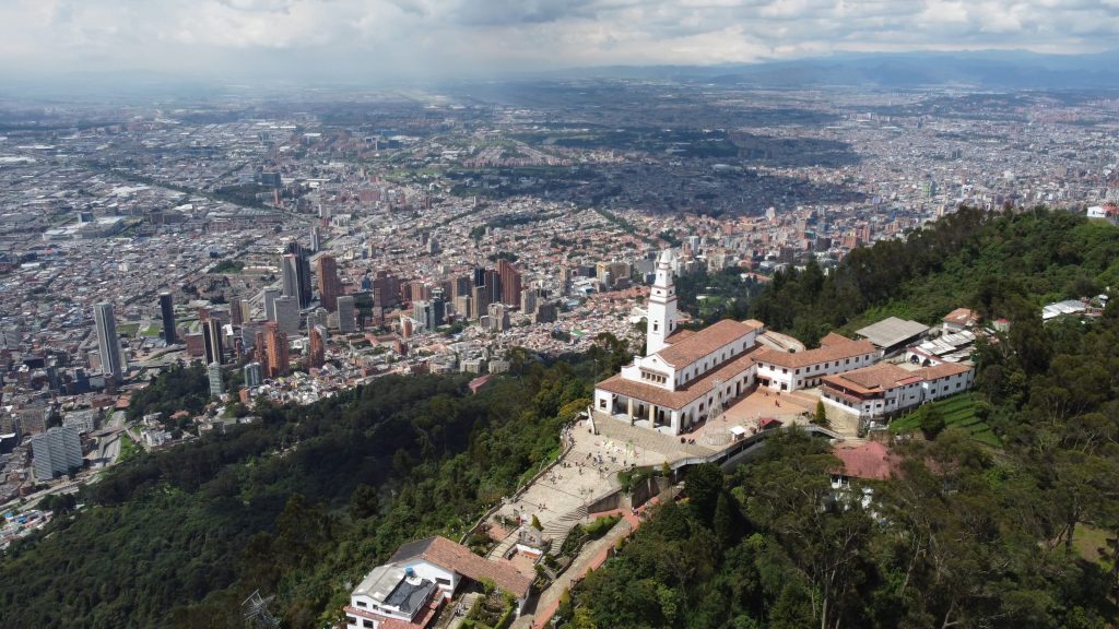 Aerial view of Monserrate Sanctuary atop a mountain overlooking Bogota, Colombia, with a sprawling cityscape under cloudy skies.