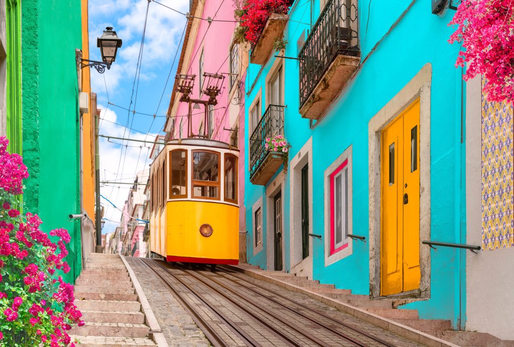 A yellow tram ascends a steep street in Lisbon, Portugal, surrounded by colorful buildings and flowers.