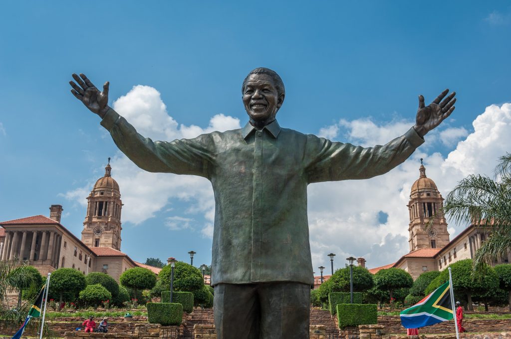 Bronze statue of Nelson Mandela with arms outstretched, in front of the Union Buildings, with South African flags.