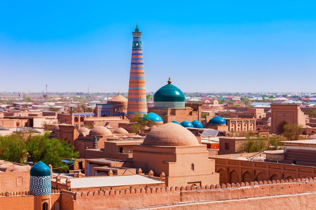 Khiva, Uzbekistan cityscape featuring historic brick buildings, blue and green domes, and a tall striped minaret under a clear blue sky.