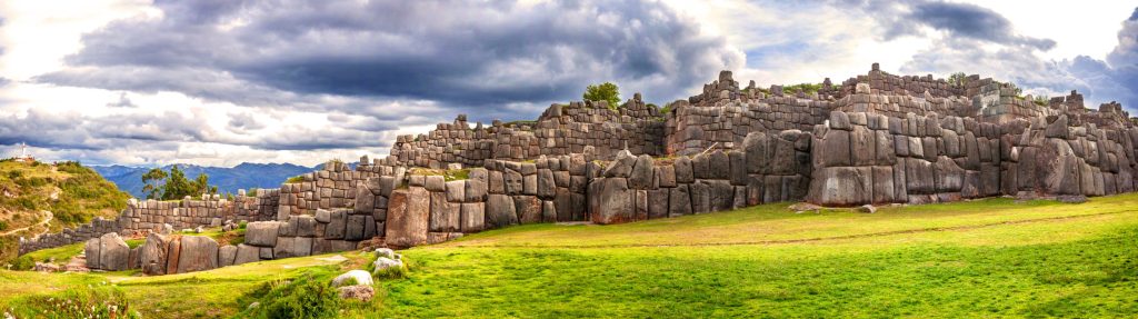 Panoramic view of Sacsayhuaman, an ancient Inca fortress in Cusco, Peru, featuring massive stone walls and green grassy foreground under cloudy skies.