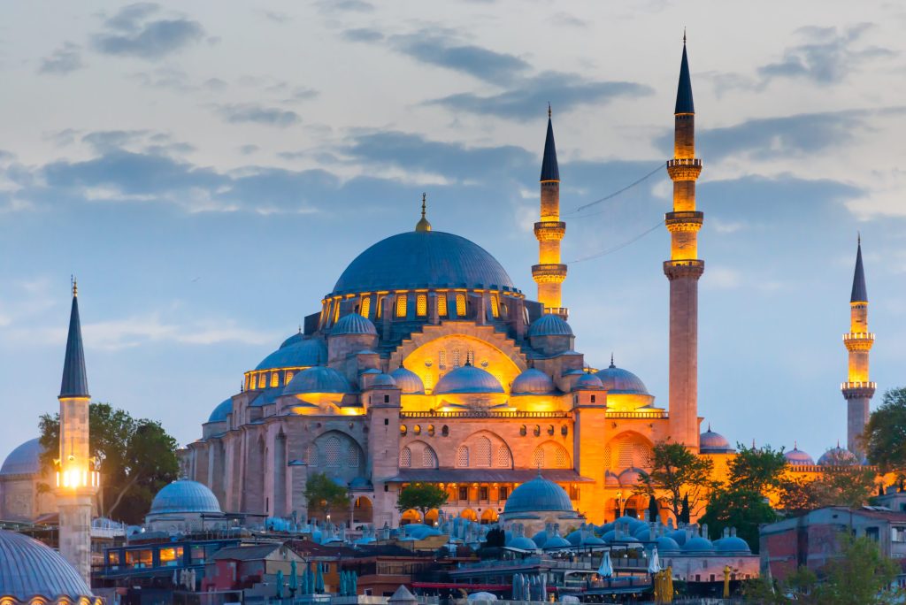 Illuminated Suleymaniye Mosque in Istanbul, Turkey at dusk. The mosque features a large dome and tall minarets against a cloudy sky.