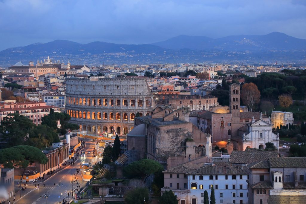 Panoramic view of the Colosseum in Rome, Italy, at dusk with city buildings and distant mountains.