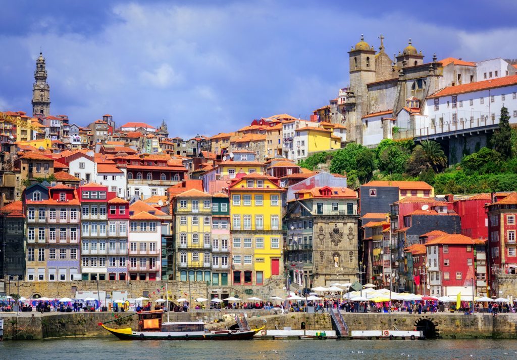 Porto, Portugal cityscape shows colorful buildings with red tile roofs along the Douro River waterfront under a blue sky.
